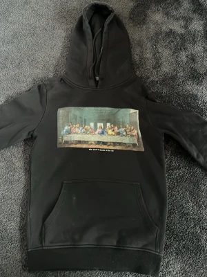 Svart hoodie med Last Supper-tryck - Cool svart hoodie med stor ficka framtill och tryck av 'The Last Supper' på bröstet. Under motivet står texten 'YOU CAN'T HANG WITH US'. Hoodien har huva med dragsko och är gjord i mjukt material, perfekt för en avslappnad streetstyle. Hoodien är ifrån Mister Tee och vita ”märket/flöcken” går bort i tvätten 