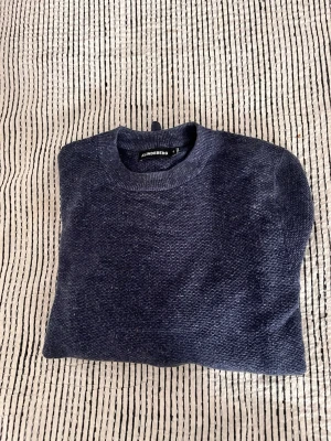 XS Mörkblå stickad tröja från J.Lindeberg - Snygg mörkblå stickad tröja från J.Lindeberg med rund hals och långa ärmar. Tröjan har en diskret struktur i stickningen och ribbade muddar vid hals, ärmslut och nederkant. Perfekt för att hålla sig varm under kyliga dagar.