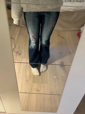 Mörkblå jeans med broderier - Snygga mörkblå jeans med rosa och vita broderier på knäna och bakfickorna. Jeansen har en klassisk bootcut-modell och coola detaljer som gör dem unika. Perfekta för dig som vill sticka ut lite extra. Materialet är denim.