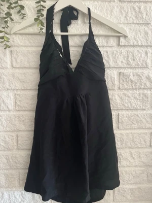 Svart halterneck linnetopp - Snygg svart halterneck linnetopp med knytning i nacken och lätt rynkade detaljer över bysten. Toppen har en luftig passform och är perfekt för en trendig look. Materialet känns lätt och svalt mot huden.