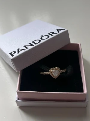 Pandora guldring med hjärtformad sten - Snygg guldfärgad ring från Pandora med en stor hjärtformad klar sten i mitten, omgiven av små gnistrande stenar längs hela ringen. Perfekt för dig som gillar romantiska och eleganta smycken. Kommer i original ask. Vill gärna få bort den så snabbt som möjligt så priset går eventuellt att få ner.💞