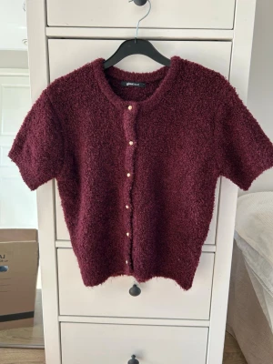 Vinröd kortärmad kofta Gina Tricot - Mysig vinröd kofta från Gina Tricot med korta ärmar och rund hals. Koftan är gjord i ett fluffigt, bouclé-liknande material och har guldfärgade knappar framtill. 