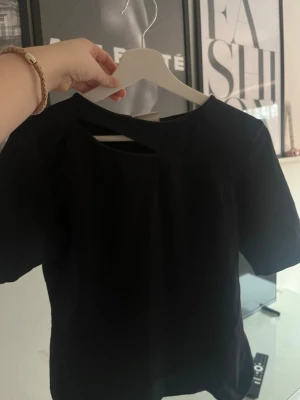 Svart topp med cut out-detalj - Säljer en svart topp med korta ärmar och en snygg cut out-detalj vid halsen. Toppen har en enkel, stilren design och är gjord i ett mjukt bomullsliknande material. Perfekt för dig som vill ha något trendigt och bekvämt.
