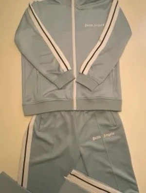 Ljusblå tracksuit från Palm Angels - Säljer en ljusblå tracksuit från Palm Angels med vita och svarta ränder längs ärmar och ben. Setet består av zip-hoodie och matchande byxor, båda med logotypen broderad framtill. Materialet är mjukt och stretchigt, perfekt för chill dagar.