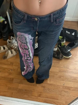 Syoersnygga liw waist baggy jeans med tryck - Unika blå jeans med ett stort graffiti-inspirerat tryck i rosa, vitt och svart längs ena benet. Jeansen har raka ben och klassisk femficksmodell. Perfekta för dig som vill sticka ut med en streetwear vibe. Rosa sömmar med hjärtan på bakfickorna. Är 170cm lång och har storlek 34. Skicka meddelande för exakta mått. 