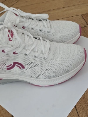Vita sneakers från Crivit  med rosa detaljer - Fräscha vita sneakers från Crivit  i 40 storlek med stickad mesh-ovandel och rosa detaljer på sidan, hälen och sulan. Skorna har vita skosnören, lågt skaft och en bekväm, vadderad insida. Perfekta för dig som gillar sportig och clean stil. Nästan nya .