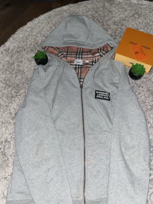 Burberry Hoodie  - Säljer en grå hoodie från Burberry med dragkedja och klassiskt rutigt foder i huvan. Märket har en svart patch med texten 'BURBERRY LONDON ENGLAND' på bröstet. Hoodien har fickor framtill och ribbade muddar. Perfekt för dig som gillar stilrena och exklusiva plagg. Dragkedjan är lite sönder men går fortfarande och dra upp enklelt ☺️🕺❤️