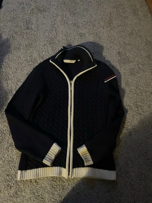 Marinblå stickad kofta med dragkedja - Snygg marinblå stickad kofta med vit dragkedja och vita ribbade muddar. Har ett diskret märke på ärmen och quiltat mönster framtill. Hög krage med vit kant och broderad detalj på axeln. Perfekt för lager på lager och stilren look. passar både xs och xss