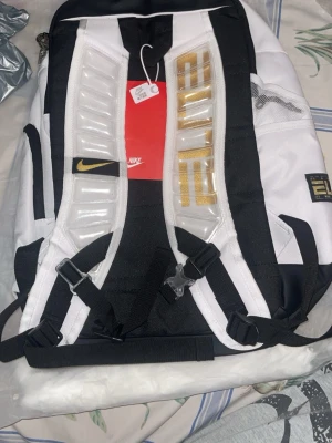 Nike ELITE vit och svart ryggsäck - Nike ELITE ryggsäck i vit och svart med gulddetaljer. Snygg design med vadderade axelband, flera fack och tydlig ELITE-logga. Tillverkad i slitstarkt material, perfekt för skola eller träning. Justerbara remmar och smarta fickor för enkel organisering.