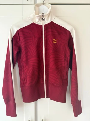 Röd och vit Puma track jacket / träningsjacka  med dragkedja - Snygg röd track jacket / träningsjacka med dragkedja från Puma med vita detaljer på axlar och krage. Tröjan har ett subtilt mönster över hela tyget och ett gult Puma-logo på bröstet. Hög krage och ribbade muddar ger en sportig look. 