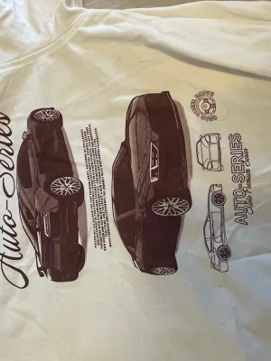 Vit t-shirt med biltryck Auto-Series - Snygg vit t-shirt med stort tryck av bilar och texten 'Auto-Series' på framsidan. T-shirten har klassisk passform och rund hals. Perfekt för dig som gillar bilar och streetstyle.