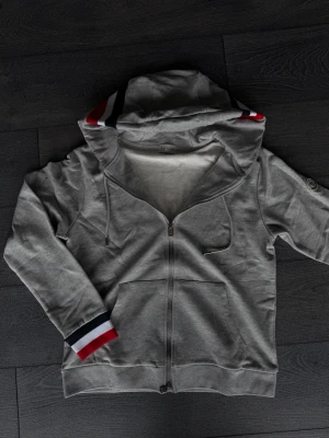Grå moncler zip up - Snygg grå zip hoodie med huva och fickor framtill. Ärmsluten och huvan har stilrena ränder i rött, vitt och blått. Tillverkad i mjukt bomullsmaterial som känns skönt mot huden. Perfekt för en avslappnad och sportig look.