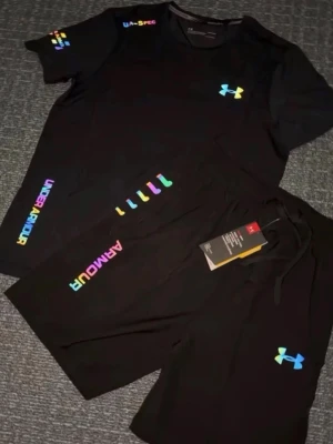 Svart träningsset från Under Armour - Snyggt svart träningsset från Under Armour med t-shirt och shorts. Båda plaggen har färgglada loggor och textdetaljer i regnbågsfärger. Perfekt för gymmet eller löprundan. Materialet är lätt och andas, vilket gör det bekvämt under träning.