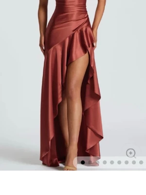 Brun asymmetrisk långklänning satin - Snygg brun långklänning i glansigt satinmaterial med omlott och hög slits framtill. Klänningen har figurnära passform och markerad midja, perfekt för dig som vill sticka ut på festen. Stilren och elegant design med volangdetaljer längs kjolen.