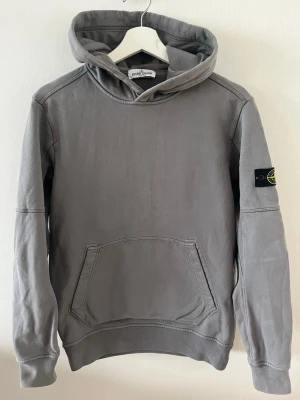 Grå hoodie från Stone Island - Snygg grå hoodie från Stone Island med klassisk huva och känguruficka framtill. Tillverkad i mjukt bomullsmaterial som är skönt att bära. Perfekt för dig som gillar stilrena och avslappnade plagg. Plagget är i mycket bra skick och passar storlek S, står junior på storleken men passar S