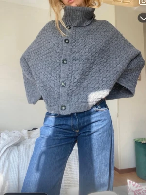 Grå stickad poncho med knappar - Säljer en grå stickad poncho med hög polokrage och stora svarta knappar framtill. Ullblandning. Funkar som en hårig jacka eller varmare kofta. 