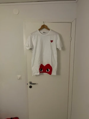 Vit Comme des Garçons Play t-shirt L - Säljer en vit t-shirt från Comme des Garçons Play i storlek L. T-shirten har korta ärmar och är gjord i mjuk bomull. På bröstet finns ett litet rött hjärta med ögon och ett stort rött hjärta med ögon längst ner på framsidan. Perfekt för dig som gillar streetwear och coola prints.