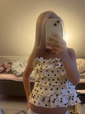 Prickig volangig tubtopp - Säljer en vit axelbandslös tubtopp med svarta prickar och flera lager volanger. Toppen är croppad och har en luftig, loose passform som ger en lekfull vibe. Perfekt för dig som vill sticka ut lite extra i sommar!, toppen funkar även som xs!!