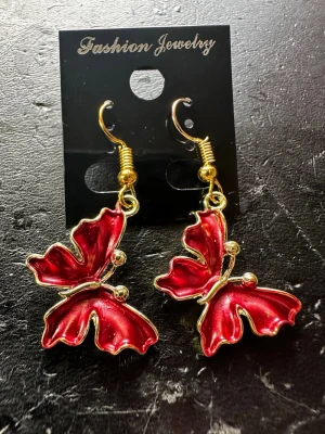 Röda fjärilsörhängen i metall - Unika örhängen i form av fjärilar med skimrande röd emalj och guldfärgade detaljer. Hängande modell med krok, perfekta för dig som vill sticka ut. Tillverkade i metall och emalj, med små guldfärgade kulor som extra detalj.