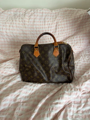 Louis Vuitton Speedy Monogram väska - Klassisk Louis Vuitton Speedy handväska i brunt monogramcanvas med detaljer i ljust brunt läder. Väskan har två handtag, guldfärgade metalldetaljer och dragkedja upptill. Ikoniskt LV-mönster över hela väskan. Perfekt för dig som gillar lyxiga accessoarer.