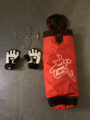 Röd boxningssäck med handskar - Säljer en röd boxningssäck med vitt kinesiskt tecken och svarta detaljer. Medföljer ett par vita och svarta fingerlösa träningshandskar samt kedja och krokar för upphängning. Perfekt för hemmaträning och kampsport.