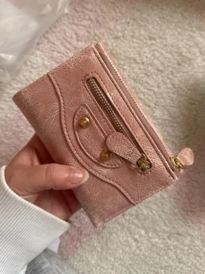 Rosa plånbok med dragkedja - Snygg rosa plånbok i skinnimitation med guldfärgade detaljer och dragkedja framtill. Plånboken har dekorativa nitar och en unik design på framsidan. Perfekt storlek för kort och kontanter.