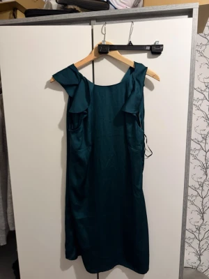 Mörkgrön klänning med volang - Snygg mörkgrön klänning i glansigt material med volangdetaljer vid axlarna och djupare ringning fram. Klänningen är ärmlös och har en rak passform som ger en chic look. Perfekt för dig som vill sticka ut lite extra.