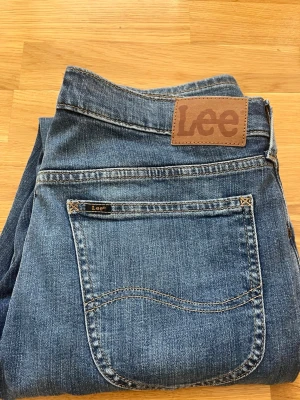 Blå jeans från Lee med rak passform - Säljer ett par klassiska blå jeans från Lee med rak passform och normal midja. Jeansen har fem fickor, tydliga sömmar och Lee-logga på bakfickan samt läderpatch i midjan. Tillverkade i slitstark denim, perfekta för en avslappnad stil. Fraktas snabbt och hör av er vid minsta funderingar!!