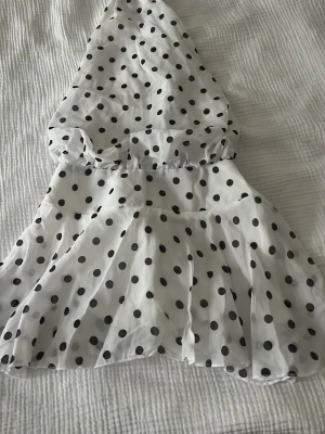 Vit prickig halterneck klänning  - Säljer en söt vit kort klänning med svarta prickar och knyt i nacken. Klänningen har en luftig kjol och är ärmlös, perfekt för varma dagar. Materialet känns lätt och flowy, troligen polyester. Supersnygg och trendig look! Klänningen är aldrig använd och är i storlek S, den är från shein. 