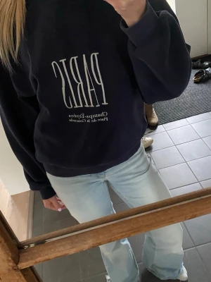 Mörkblå sweatshirt med Paris-tryck - Säljer en mörkblå sweatshirt med stort vitt tryck där det står 'PARIS' på bröstet och mindre text under. Tröjan har rund hals, långa ärmar och ribbade muddar vid ärmslut och nederkant. Perfekt för en avslappnad och trendig look.