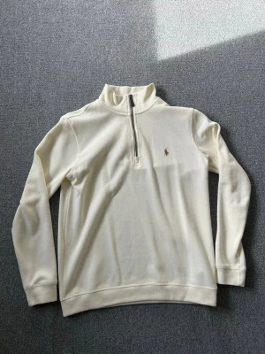 Beige halvzip från Polo Ralph Lauren - Klassisk vit långärmad tröja från Polo Ralph Lauren med halv dragkedja och broderad logga på bröstet. Tröjan har hög krage och ribbade muddar. Perfekt för en clean och stilren look.