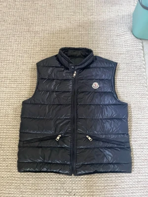 Navy blå dunväst från Moncler - Snygg svart dunväst från Moncler med hög krage och två dragkedjefickor framtill. Västen har quiltad design och ikonisk Moncler-logga på bröstet. Perfekt för lager-på-lager och håller dig varm med stil. Materialet är glansigt och lätt.