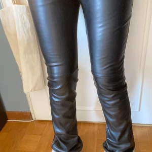 Svarta bootcut byxor i skinnimitation - Säljer ett par svarta byxor i fuskskinn med bootcut-modell och hög midja. Byxorna har en glansig finish och sitter tight över låren för en trendig och edgy look. Från Lindex. Har inga fickor och är högmidjad. Säljs troligtvis inte längre. Bara använd en gång 