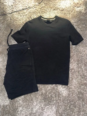 Svart set från BOSS med t-shirt och shorts - Säljer ett svart set från BOSS som består av en kortärmad t-shirt och matchande shorts med dragsko i midjan. Båda plaggen har diskret BOSS-logga broderad framtill. Materialet känns mjukt och bekvämt, perfekt för en avslappnad stil.