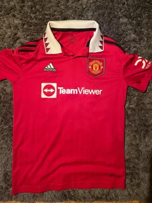 Manchester United matchtröja Adidas - Säljer en röd Manchester United matchtröja från Adidas med vit krage och svarta detaljer. Tröjan har klubbmärke på bröstet, TeamViewer sponsortryck och klassiska Adidas-ränder på axlarna. Perfekt för fotbollsfans som vill visa sitt lagstöd.