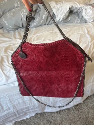 Röd as snygg väska - Säljer en snygg röd väska med ormmönstrad struktur. Väskan har silverfärgade kedjehandtag och dekorativa sömmar längs kanterna. Invändigt finns svart foder och innerfack med dragkedja. Köpt på Plick 