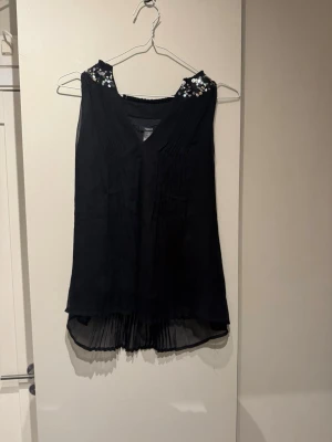 Svart halterneck topp med paljetter - Snygg svart halterneck topp med v-ringning och stora silvriga paljetter på axlarna. Toppen har ett lätt transparent, plisserat tyg nedtill och en flowy passform. Perfekt för dig som vill sticka ut lite extra.