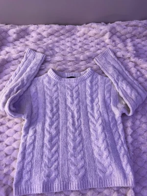 Ljuslila kabelstickad tröja - Mysig ljuslila kabelstickad tröja med rund hals och långa ärmar. Tröjan har ett klassiskt flätmönster och ribbade muddar vid ärmslut och nederkant. Perfekt för kyliga dagar och enkel att matcha med jeans eller kjol.