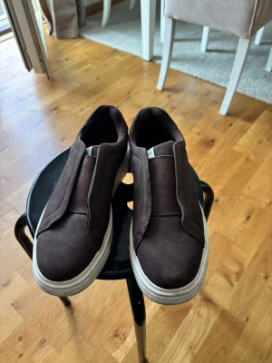 Bruna slip-on sneakers med vit sula - Mörkbruna slip-on sneakers med tjock vit sula och minimalistisk design. Ovandelen är i textilmaterial och skon saknar snörning för en clean look. Perfekta för dig som gillar enkel och stilren streetstil.