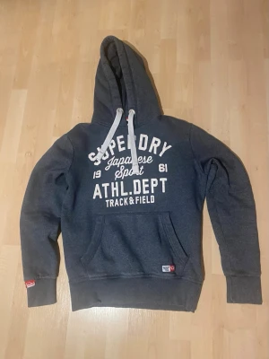Blå hoodie från Superdry - Blå hoodie från Superdry med vit text på bröstet och stora snören i huvan. Klassisk känguruficka framtill och ribbade muddar. Materialet är mjukt och bekvämt, perfekt för en avslappnad look. Snyggt tryck med ATHL.DEPT och Track & Field.