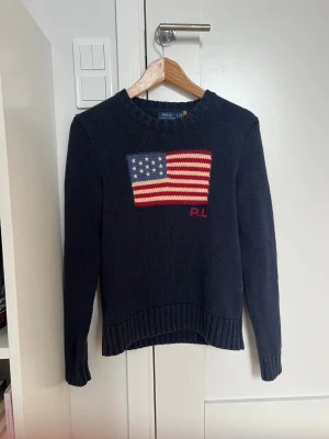 Mörkblå stickad tröja Polo Ralph Lauren - Mörkblå stickad tröja från Polo Ralph Lauren med amerikansk flagga i rött, vitt och blått på bröstet samt RL-broderi. Den är bara lite krympt. Tröjan har rund hals, långa ärmar och ribbstickade muddar vid ärmslut och nederkant. Perfekt för dig som gillar klassisk stil. Den är i fantastiskt skick och priset är inte hugget i sten! 
