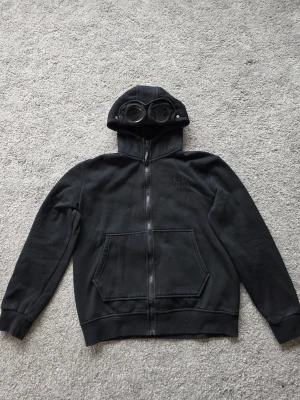 Svart hoodie med goggles från C.P. Company - Cool svart hoodie från C.P. Company med unika goggles integrerade i huvan. Tröjan har dragkedja, två stora fickor framtill och ribbade muddar. Materialet är mjukt och bekvämt, perfekt för dig som vill sticka ut med en streetwear-look.