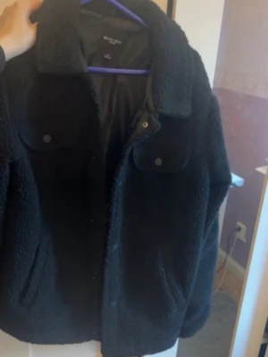 Svart teddyjacka från Brave Soul - Mysig svart teddyjacka från Brave Soul med klassisk krage och två bröstfickor med knappar. Jackan har en fluffig och mjuk textur, perfekt för att hålla dig varm. Stängs med knappar framtill och har en avslappnad passform.