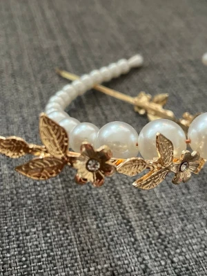 Diadem med pärlor och guldfärgade blommor - Snyggt diadem dekorerat med stora vita pärlor och guldfärgade metallblommor och blad. Blommorna har små glittrande stenar i mitten och diademet har en elegant, romantisk känsla. Perfekt accessoar för att lyfta din look med en touch av lyx.
