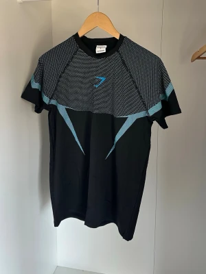 Svart Gymshark t-shirt med blå detaljer - Säljer en svart t-shirt från Gymshark med blå geometriska mönster och logga på bröstet. Tröjan har korta ärmar och är tillverkad i ett lätt och stretchigt material som passar perfekt för träning och sportaktiviteter. OBS rep. passar S som muselfit och passar även M fast lite tajtare 