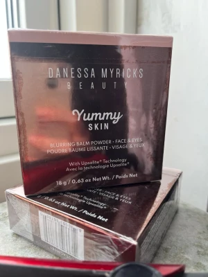 Danessa Myricks Yummy Skin Balm - Yummy Skin Blurring Balm Powder från Danessa Myricks Beauty i nyansen 3,75. Kommer i en rund, kopparfärgad dosa med spegel och lock med logga. Formulan är krämig och puderliknande, perfekt för att jämna ut hudtonen och ge en naturlig finish.