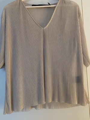 Beige ribbad v-ringad topp - Luftig beige topp med v-ringning och ribbad struktur. Toppen har korta ärmar och en lös passform som ger en avslappnad look. Perfekt att matcha med jeans eller kjol för en enkel stil.