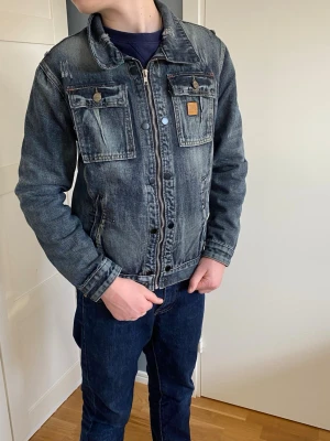 Blå jeansjacka från Burberry - Snygg blå jeansjacka från Burberry med klassisk passform. Jackan har två bröstfickor med lock och knappar, samt två snedställda sidofickor. Detaljer i brunt läder med Burberry London England-märke på bröstet. Dragkedja framtill och kontrastsömmar.