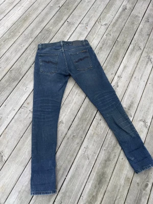 Mörkblå slim fit jeans från Nudie - Säljer ett par mörkblå slim fit jeans från Nudie Jeans med klassiska fem fickor och snygga slitningar på låren. Jeansen har kontrastsömmar och Nudie-logga på bakfickan. Perfekta för dig som gillar en smal passform och tidlös stil.