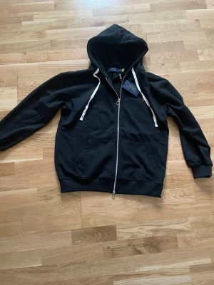 Svart hoodie från Polo Ralph Lauren - Svart hoodie med dragkedja från Polo Ralph Lauren. Klassisk design med huva och vita dragsnören, samt röd broderad logga på bröstet. Mjuk bomullskänsla och känguruficka framtill. Perfekt för en avslappnad och stilren look.